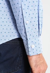 Camisa de botones de color azul claro con pequeño patrón floral en azul oscuro, que presenta botones blancos en el puño y un corte estructurado.
