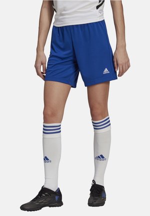 Osoba ubrana w niebieskie spodenki Adidas, białe podkolanówki z niebieskimi paskami i czarne korki piłkarskie Adidas na jednolitym tle.
