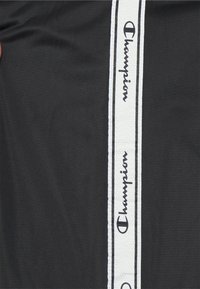 Tissu noir avec une bande blanche verticale présentant des logos noirs Champion répétés et du texte.