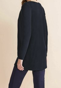 Mörkblå långärmad tunika i mjukt tyg, med rundad halsringning och en något kurvig nederkant, kombinerad med mörka byxor.