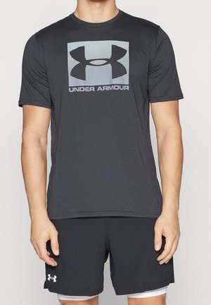 Mężczyzna ubrany w ciemnoszarą koszulkę Under Armour z dużym logo i pasujące szorty z małym logo na lewej nodze.