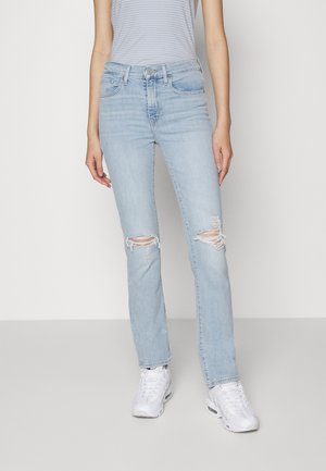 Vrouw in lichtblauwe gescheurde spijkerbroek, witte sneakers en een mouwloze grijze gestreepte top, staand tegen een effen lichte achtergrond.