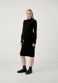 Nėščia moteris vilki juodą ilgo rankovės vidutinio ilgio suknelę su užrišama juosta juosmenyje, juodas permatomus taškuotus kojines ir juodus loaferius.