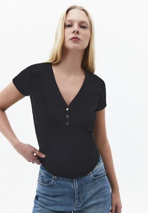 Jonge vrouw met lang blond haar, gekleed in een zwart V-hals T-shirt met korte mouwen en knopen, en een hooggetailleerde blauwe spijkerbroek, poserend tegen een witte achtergrond.