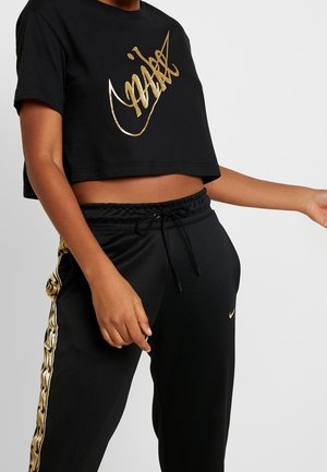 Personne portant un t-shirt court noir avec le logo Nike doré et un pantalon noir avec une bande ondulée dorée et un petit swoosh Nike sur la jambe gauche.