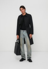 Zwarte lange jas over een zwart T-shirt, vervaagde blauwe jeans en zwarte schoenen. Houdt een zwarte totebag vast. Modern ontwerp, op maat gemaakt.