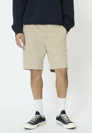 Beige Baumwollshorts mit lockerem Sitz, seitlichen Taschen und einem sauberen Saum. Kombiniert mit schwarzen Turnschuhen und weißen Socken.