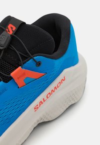 Baskets de sport bleues avec une tige texturée, des détails en maille noire, une touche orange, et une semelle blanche proéminente avec des motifs d'adhérence.