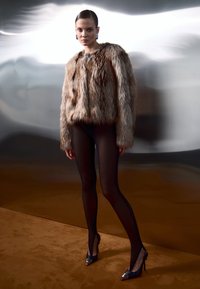 Chaqueta de piel sintética en tonos marrones, con textura gruesa y diseño oversize. Combinada con medias negras transparentes y tacones negros con detalles plateados.