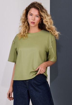 Kvinde med krøllet blondt hår iført en olivengrøn top med korte ærmer og mørkeblå jeans, stående foran en tofavet grå baggrund.