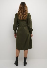 Zeleno-olivové šaty s dlhými nariasenými rukávmi, priliehavým korzetom a sukňou vo forme písmena A, vyrobené z hladkej látky, doplnené čiernymi členkovými čižmami.