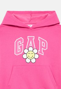 Sweatshirt de algodão rosa com o logótipo branco "GAP" e uma ilustração de margarida sorridente ao centro, com um bolso tipo canguru na frente.