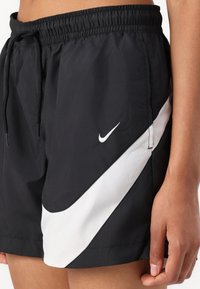 Melnas Nike peldbikses ar baltu diagonālu svītru, elastīgu jostu, ar auklu aizdari un logo detaļu kreisajā pusē.