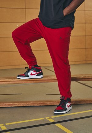 Person, die rote Jogginghosen und schwarze, rote und weiße Nike-Sneakers trägt, steht mit einem Fuß auf einer Holzbank in einer Sporthalle.
