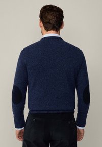 Maglione lavorato a maglia blu navy con finitura testurizzata, caratterizzato da toppe nere sui gomiti e da un colletto e polsini a costine. Pantaloni scuri visibili sottostante.