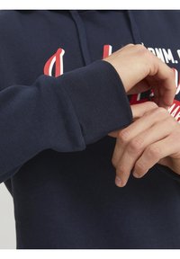 Námořnická modrá mikina s kapucí, která má ribbené man�žety, textilní texturu a tučné červené písmo na hrudi. Materiál vypadá měkký a pohodlný.