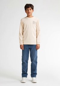 Natürlich beiges Langarm-T-Shirt mit kleinem Aufdruck auf der Brust, kombiniert mit blauen Denim-Jeans und weißen Sneakers, vor neutralem Hintergrund stehend.