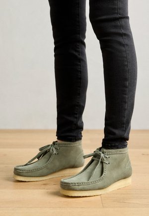 Grüne Wildleder-Stiefeletten mit weicher Sohle, die ein Schnürdesign und gestickte Details entlang der Kanten aufweisen. Tragen zu schwarzen Jeans.
