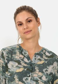 Grøn blomstret bluse med en rund krave, der har en knaplukning, lyseblå og gule accenter, samt et blødt, tekstureret stof.