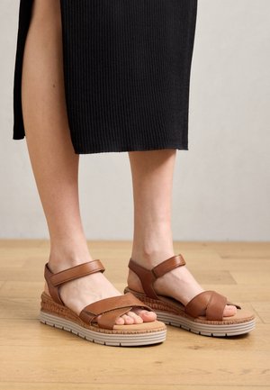 Person iført brune sandaler med brede hvide såler og en sort ribstrikket nederdel, står indendørs på et lyst trægulv.