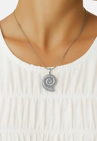 Sølvved pendant i form av en spiral skjell på en tynn kjede, båret over en hvit, rynket stofftopp. Teksturert, metallisk finish.
