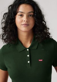 Donkergroene polo shirt gemaakt van textured stof, met een kraag en vier witte knopen, en een klein rood Levi's logo patch op de borst.