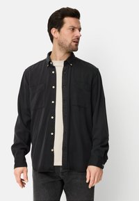 Chemise noire boutonnée en coton, avec deux poches poitrine et des boutons blancs. Portée sur un t-shirt clair, associée à un jean foncé.