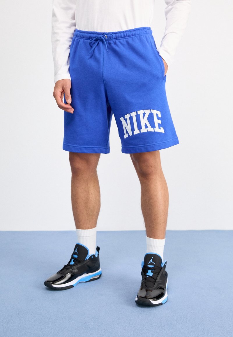 Niebieskie spodenki wykonane z mieszanki bawełny, z elastycznym pasem ze sznurkiem i białym logo "NIKE" na lewym udzie. Czarne sneakersy z niebieskimi akcentami.