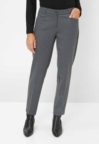 Vrouw in grijze gestreepte rechte broek, zwart lange mouwen top en zwarte enkellaarzen, met een hand in de zak.