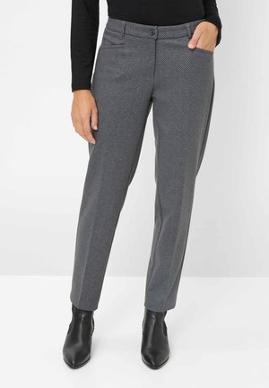 Kvinde iført grå mønstrede lige ben bukser, sort langærmet top og sorte ankelstøvler, med den ene hånd i lommen.