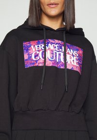 Černá bavlněná mikina s širokým, barevným grafickým potiskem, který obsahuje text "VERSACE JEANS COUTURE". Navržena s kapucí na šňůrku a žebrovaným lemem.