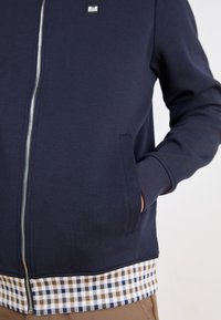 Navy zip-up sweatshirt with a checkered bottom hem in brown, white, and blue. Features side pockets and a square logo patch on the chest.