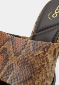 Približen posnetek zaponke čevlja z vzorcem rjave in bež kačje kože nad črnim oblazinjenim vložkom z zlatim logotipom.