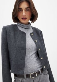 Harmaa teksturoitu lyhyt takki, jossa on kultaiset napit, puettuna harmaan poolopaidan päällä. Ominaisuuksiin kuuluu siisti kaula-aukko ja etutaskut.