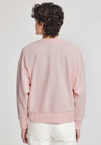 Felpa rosa chiaro con scollo a giro, spalle basse e polsini a costine. Tessuto morbido, orlo rifinito e vestibilità comoda. Pantaloni bianchi visibili.