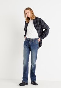 Camisa preta de padrão xadrez de mangas longas sobre uma t-shirt branca, combinada com jeans azul claro e sapatos pretos. Estilo casual, corte reto.