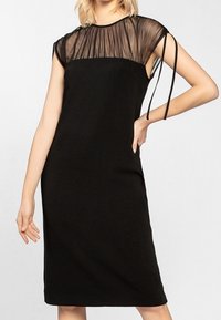 Černé šaty s průhledným vrškem se strukturou skládání, krátkými rukávy a nastavitelnými ramínky. Těsný střih s délkou ke kolenům.