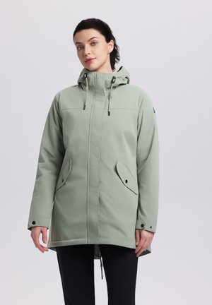 Frau trägt eine hellgrüne Winterjacke mit Kapuze und Vorderentaschen sowie schwarze Hose, steht vor einem schlichten hellen Hintergrund.