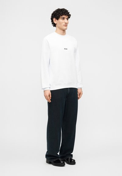 Homme aux cheveux bouclés portant un sweat-shirt blanc MSGM, un jean ample foncé, et des chaussures noires brillantes, debout devant un fond clair uni.