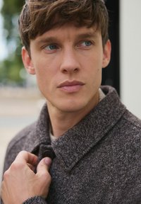 Noor mees siniste silmade ja pruunide juustega kinnitab välistingimustes oma tumeda halli tekstuuriga jope nööbi.