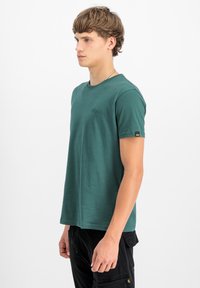 Zelené tričko s krátkým rukávem vyrobené z měkké tkaniny má kulatý výstřih, decentní logo na hrudi a značkovou etiketu na rukávu.