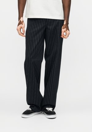 Oseba, ki nosi široke hlače s črnimi črtami, črno-bele superge in belo srajco, stoji z rokami sproščeno ob straneh.