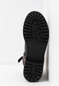 Semelle de botte noire avec des crampons profonds présentant des motifs en croix et en ligne, montrant partiellement la tige à imprimé léopard et une fermeture éclair sur fond blanc.