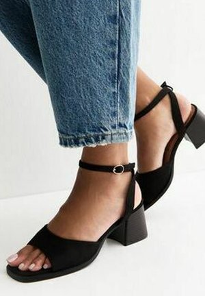Pies con sandalias negras de tacón grueso y tiras en el tobillo, combinadas con jeans azules cortos de mezclilla con bajos deshilachados.