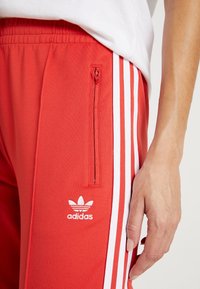 Červené sportovní kalhoty s bílými bočními pruhy, obsahující zipovou kapsu a vyšitým logem. Hladká tkanina se strečovým střihem.