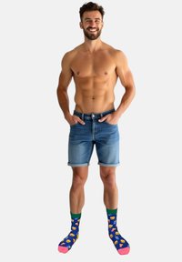 Denimshorts med opkrogede kanter i lyseblå farve. Farverige sokker med madmønstre på en mørkeblå baggrund. Mand stående med hænderne i lommerne.