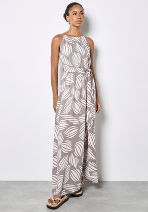 Kobieta stojąca w szarej, bezrękawnikowej sukience maxi z dużymi białymi wzorami liści i paskiem w talii, w połączeniu z beżowymi sandałami na platformie.
