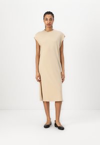 Robe midi beige avec manches courtes, fente sur le côté, tissu doux, silhouette décontractée, associée à des chaussures plates noires. Design minimaliste.