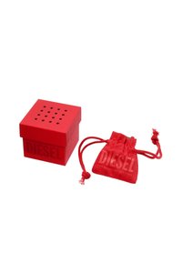 Punainen neliömäinen laatikko, jossa on teksturoitu pinta ja pieniä reikiä päällä, ja jossa lukee "DIESEL". Mukana on myös matching punainen kangaspussi, jossa on nyöri.