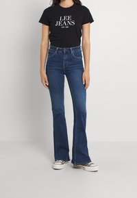 Svart t-shirt med texten "LEE JEANS EST. 1889", högmidjade blå flare-jeans och mönstrade sneakers. Slätt tyg och klassisk passform.